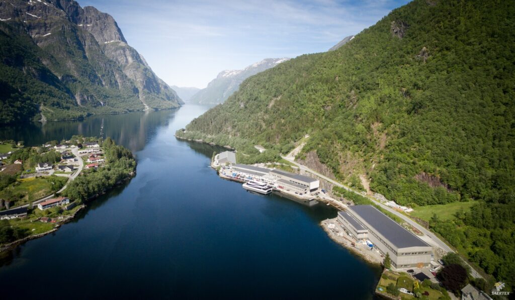 Luftaufnahme eines tiefen Fjords zwischen grünen Bergen mit einem kleinen Ort und Hafenanlagen am Ufer