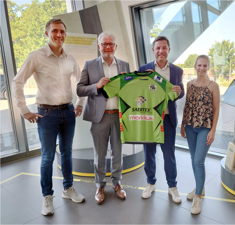 Vier Personen stehen in einem hellen Raum und halten gemeinsam ein grünes Sporttrikot mit Sponsorenlogos.