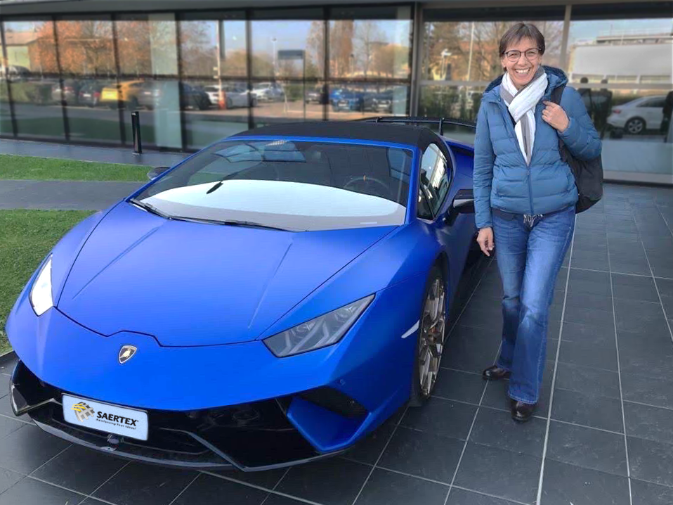 Eine lächelnde Frau steht neben einem blauen Lamborghini vor einem modernen Gebäude.