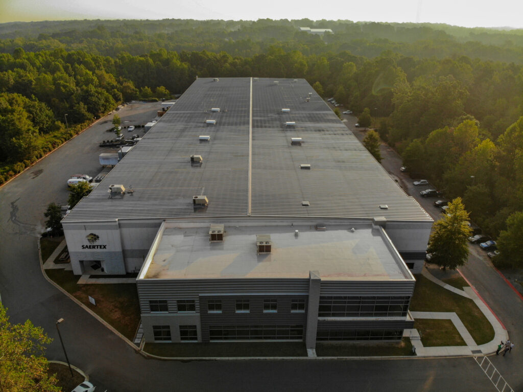 Luftbild eines großen Industriegebäudes mit Metalldach, umgeben von Wald und Parkplätzen.