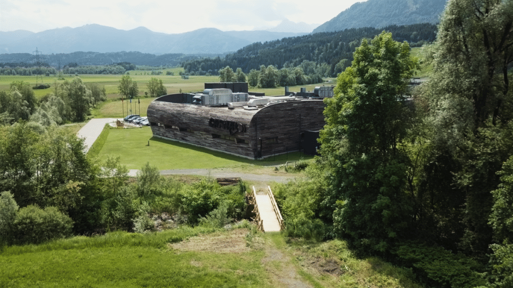 Ein großes, modern geformtes Holzgebäude steht in einer grünen Landschaft mit Bäumen, Wiesen und Bergen im Hintergrund.