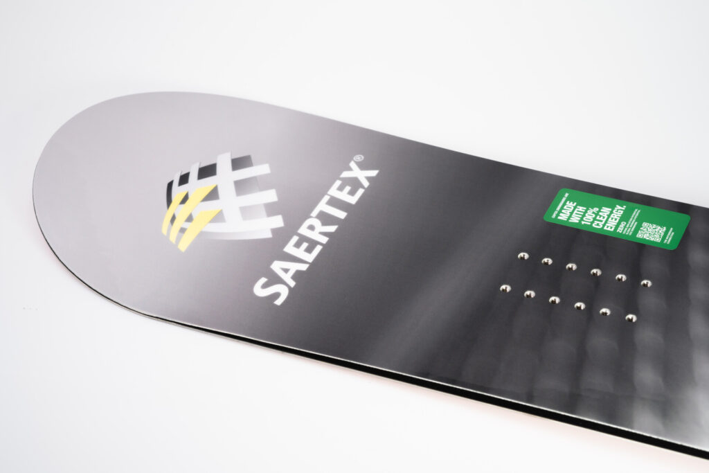 Ein schwarzes Snowboard mit SRTEX-Logo und grünem Aufkleber auf hellem Hintergrund.