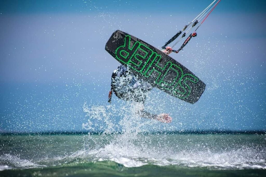 Ein Kitesurfer springt hoch über dem Wasser, während das Board mit dem Schriftzug „SHREDS“ sichtbar ist.