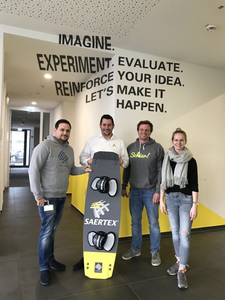 Vier Personen stehen in einem Flur und halten ein Wakeboard mit der Aufschrift SAERTEX.