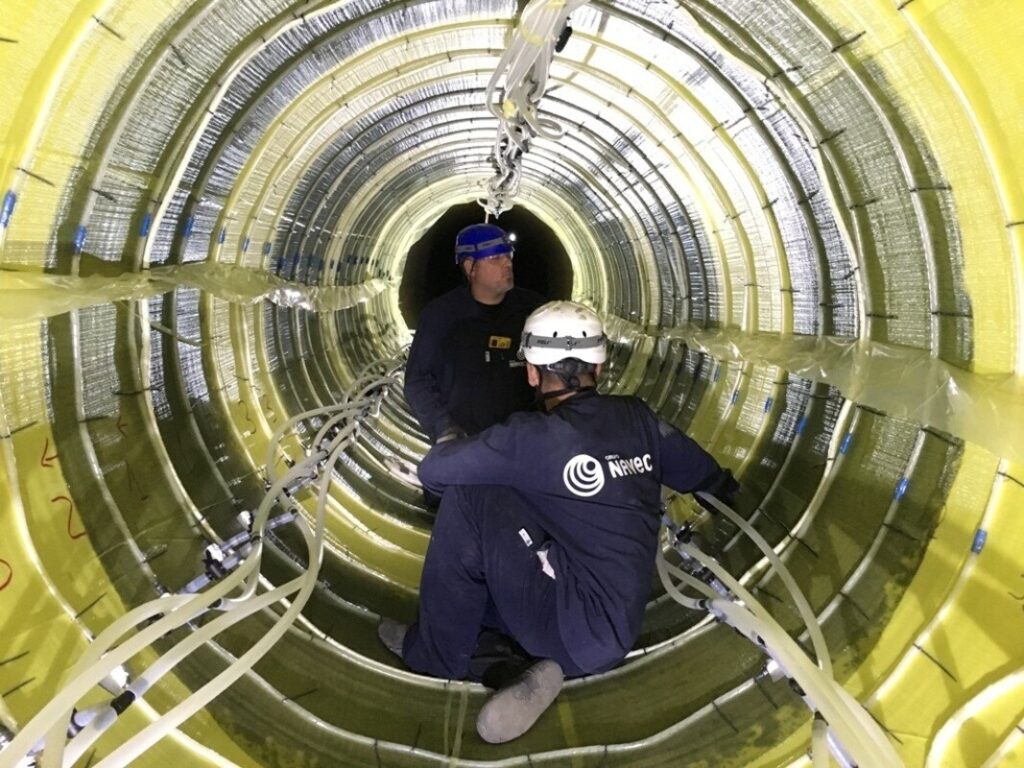 Zwei Arbeiter mit Helmen sitzen in einem mit gelber Isolierung ausgekleideten Tunnel und verlegen Leitungen.