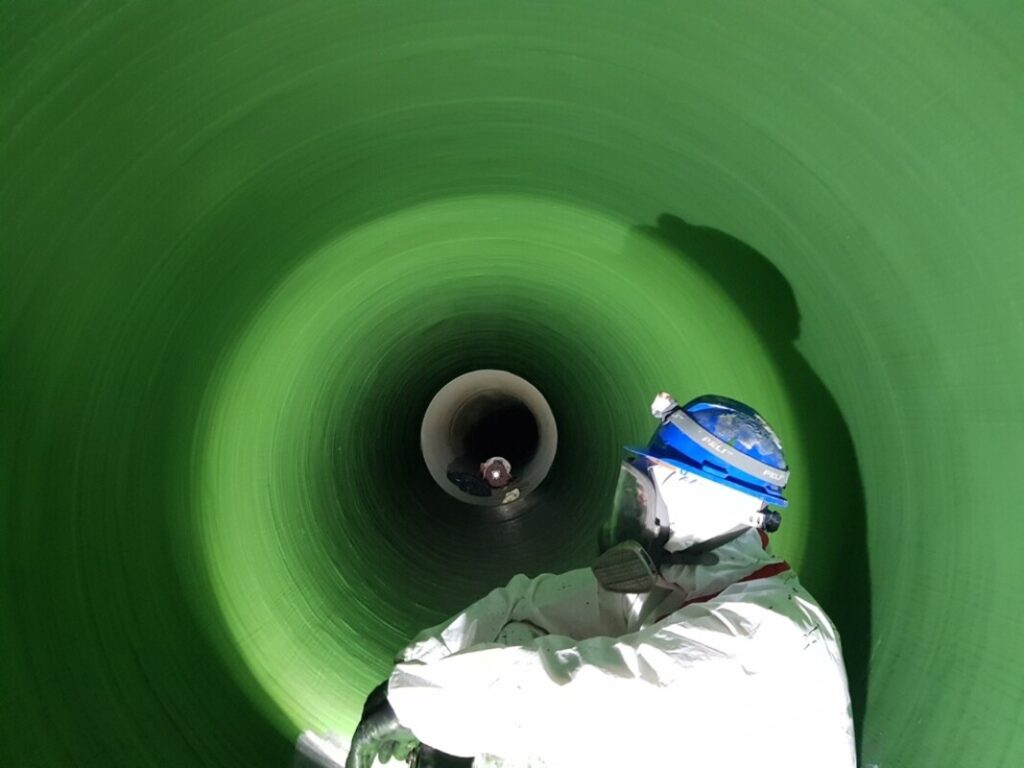 Eine Person mit Helm und Schutzanzug sitzt in einem großen grünen Rohr oder Tunnel.