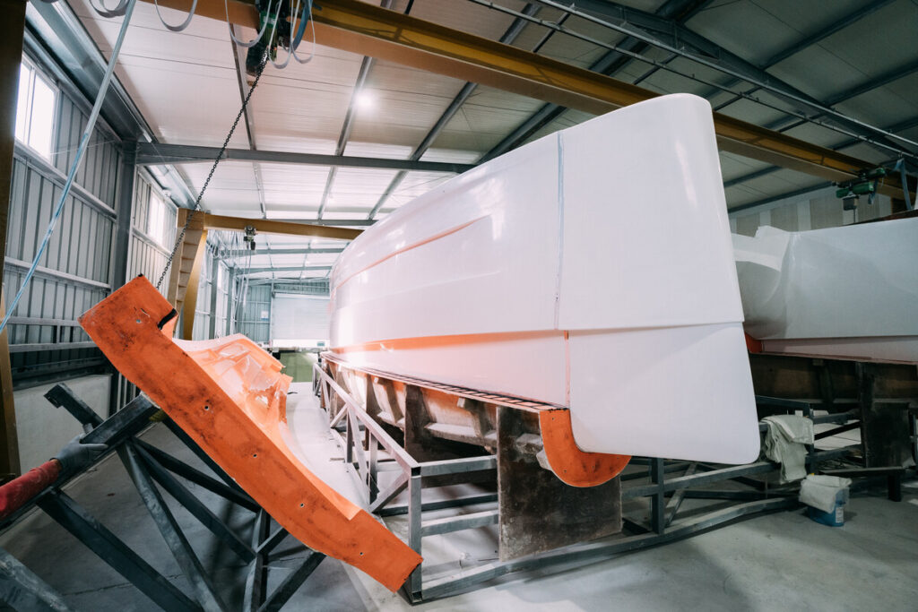 Ein großes weißes Bootsrumpf- oder Schiffsteil in einer industriellen Werkhalle mit Metallkonstruktionen und orangefarbenen Formteilen.