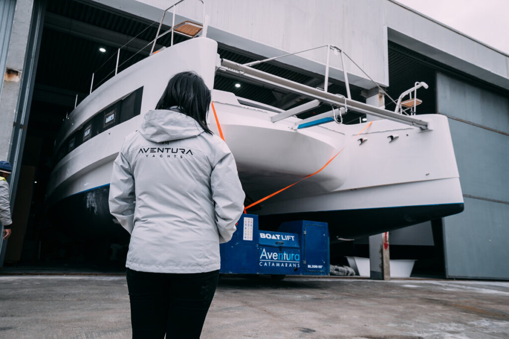Eine Person im weißen Aventura-Yachts-Jacken steht vor einem großen Katamaran in einer Werft- oder Hallenumgebung.