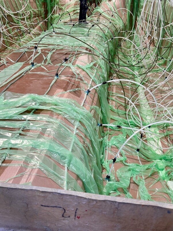 Nahaufnahme einer mit grünem Folienmaterial umwickelten technischen Anlage mit zahlreichen Schläuchen und Kabeln.