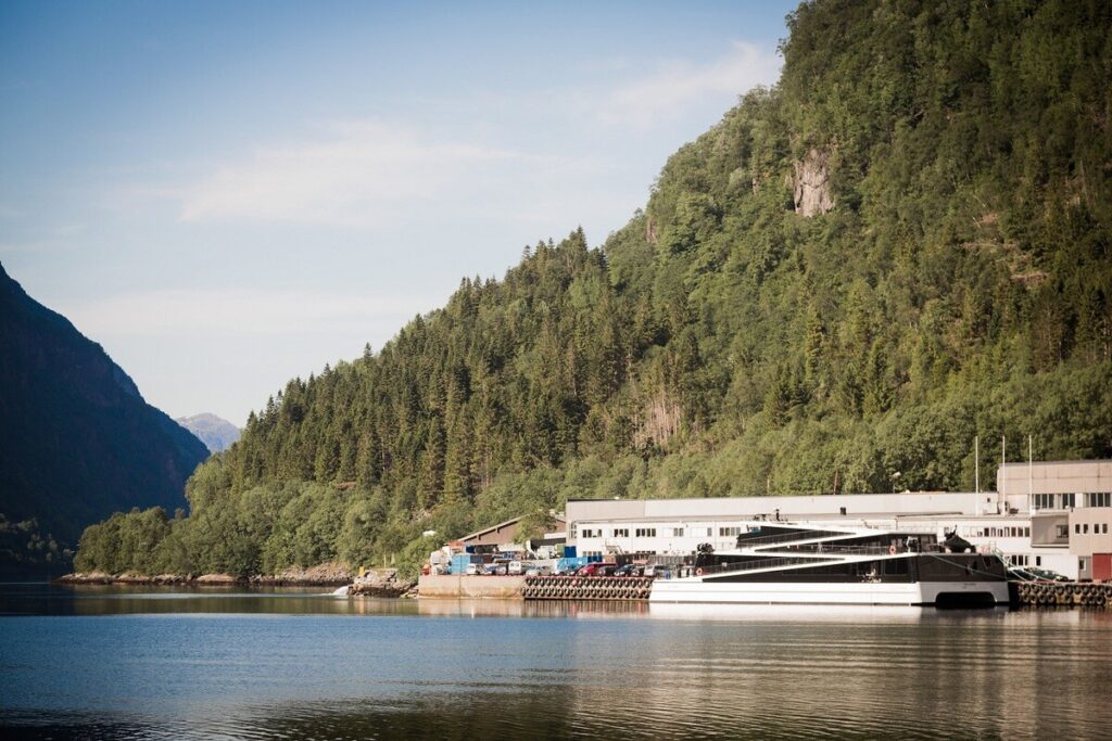 Ein Schiff liegt an einem Ufer mit Gebäuden vor bewaldeten Bergen und ruhigem Wasser.