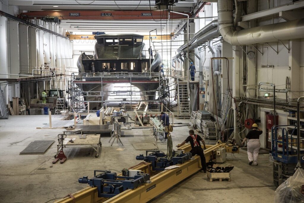 Ein großes Schiff in einer industriellen Werfthalle, umgeben von Arbeitern, Gerüsten und Maschinen.
