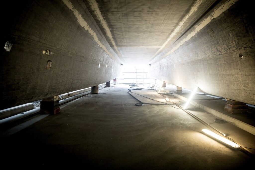 Blick durch einen dunklen Betonraum mit hellem Licht am Ende und Baugeräten am Boden.