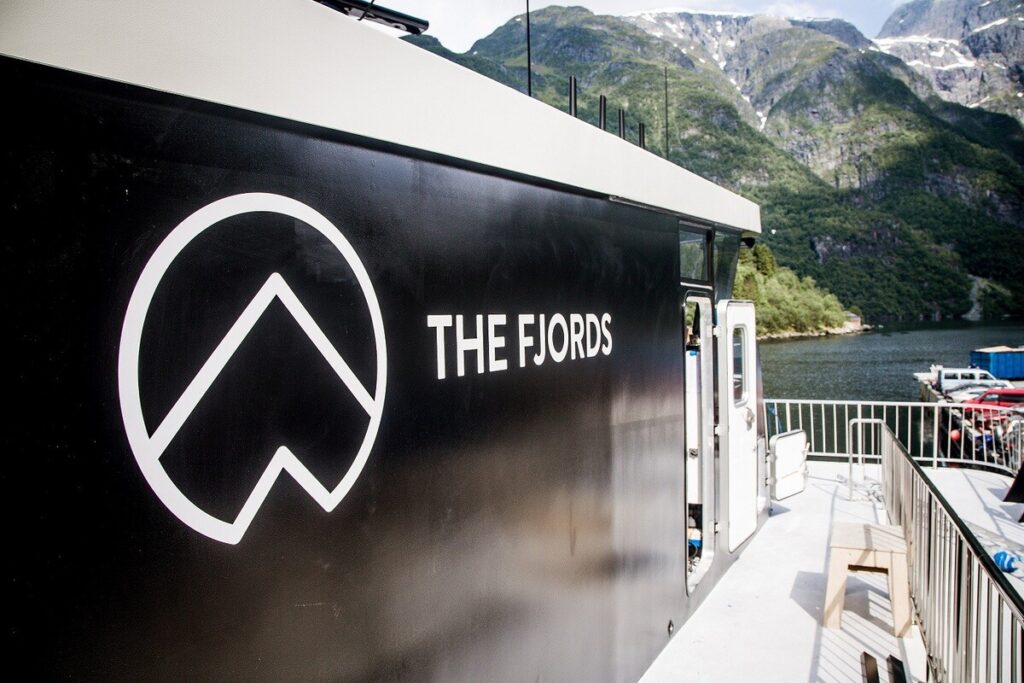Ein schwarzes Boot mit dem Schriftzug „THE FJORDS“ liegt vor steilen Bergen und einem Fjord.