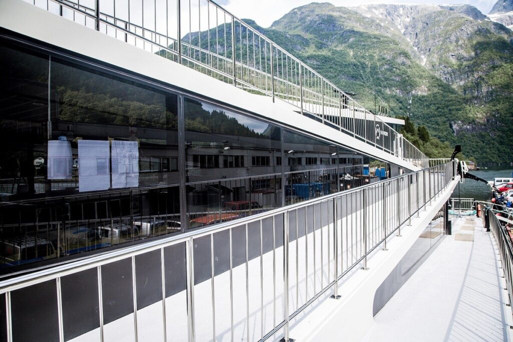 Moderne Glasfassade eines Gebäudes an einer Promenade mit Geländern, im Hintergrund Berge und Wasser.