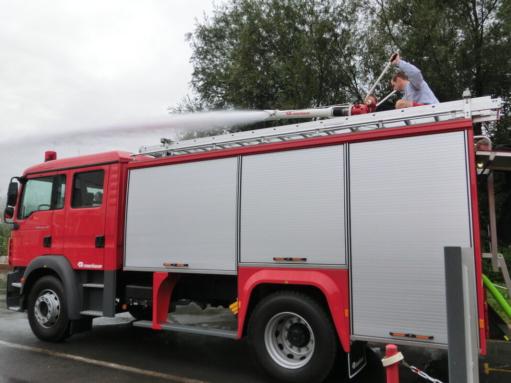 Ein rotes Feuerwehrfahrzeug löscht mit einem Wasserstrahl, während ein Feuerwehrmann oben am Schlauch arbeitet.