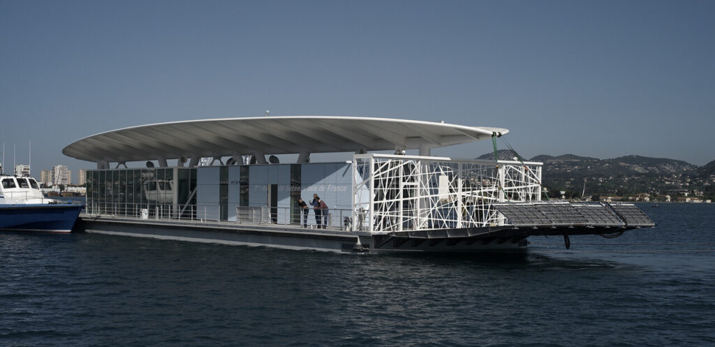Eine moderne schwimmende Plattform mit weißem Dach liegt auf dem Meer, mit einem Boot daneben und Küste im Hintergrund.