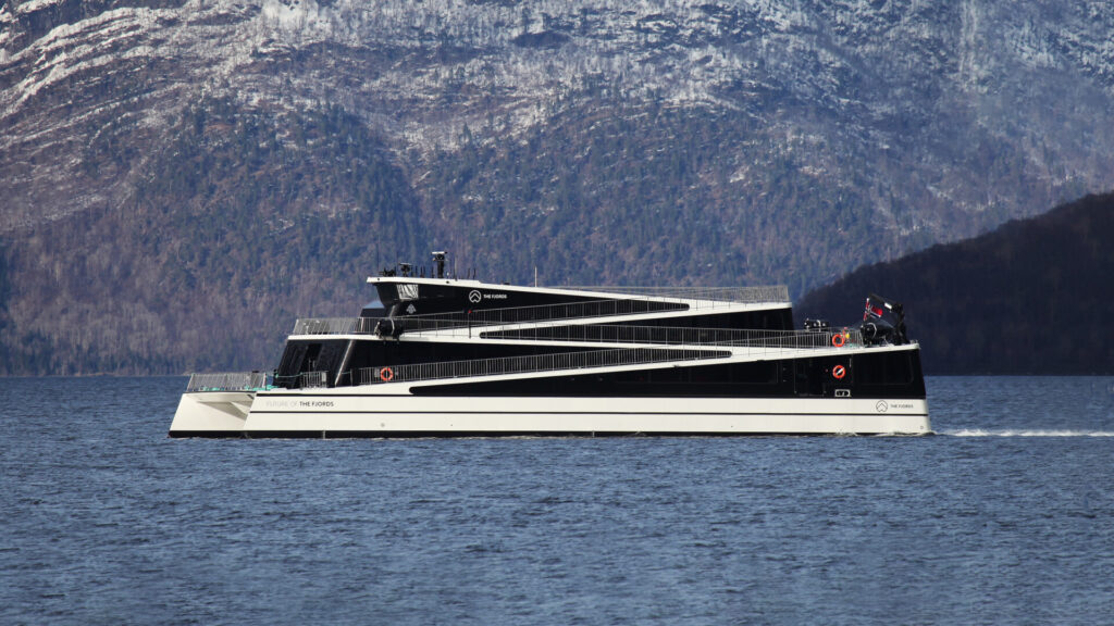 Ein modernes schwarz-weißes Passagierschiff fährt über ruhiges Wasser vor einer steilen, schneebedeckten Bergkulisse.