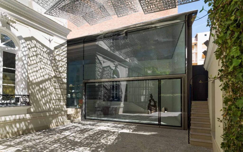 Ein moderner Glasbau in einem sonnigen Innenhof mit Musterschatten auf den Wänden und dem Boden.
