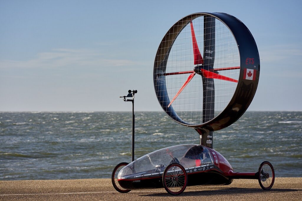 Ein ungewöhnliches, dreirädriges Windfahrzeug mit großem, rundem Rotor steht an der Küste vor dem Meer.