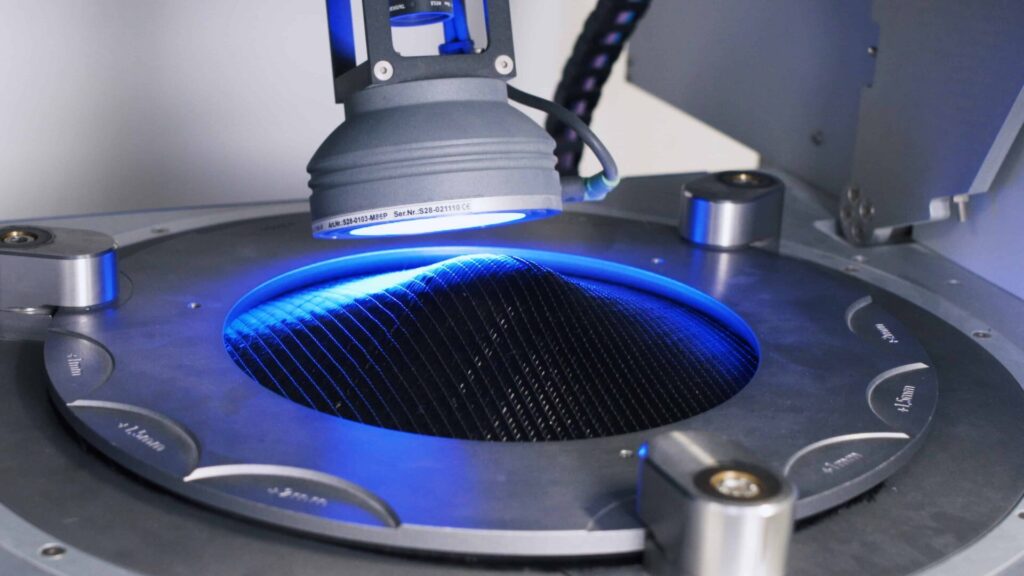 Eine Maschine beleuchtet ein rundes, dunkel schimmerndes Objekt mit blauem Licht in einer technischen Umgebung.