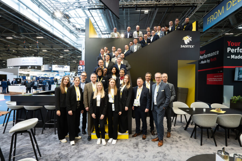 Eine große Gruppe von Menschen posiert auf einem Messestand vor einer schwarzen Treppe mit dem Logo SAERTEX.
