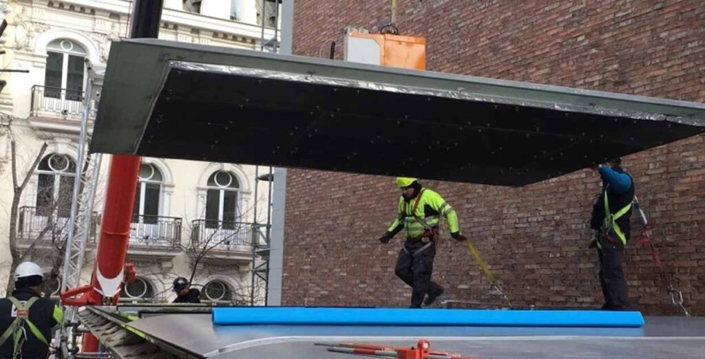 Mehrere Arbeiter heben und befestigen eine große, dunkle Überdachungsplatte auf einer Baustelle.