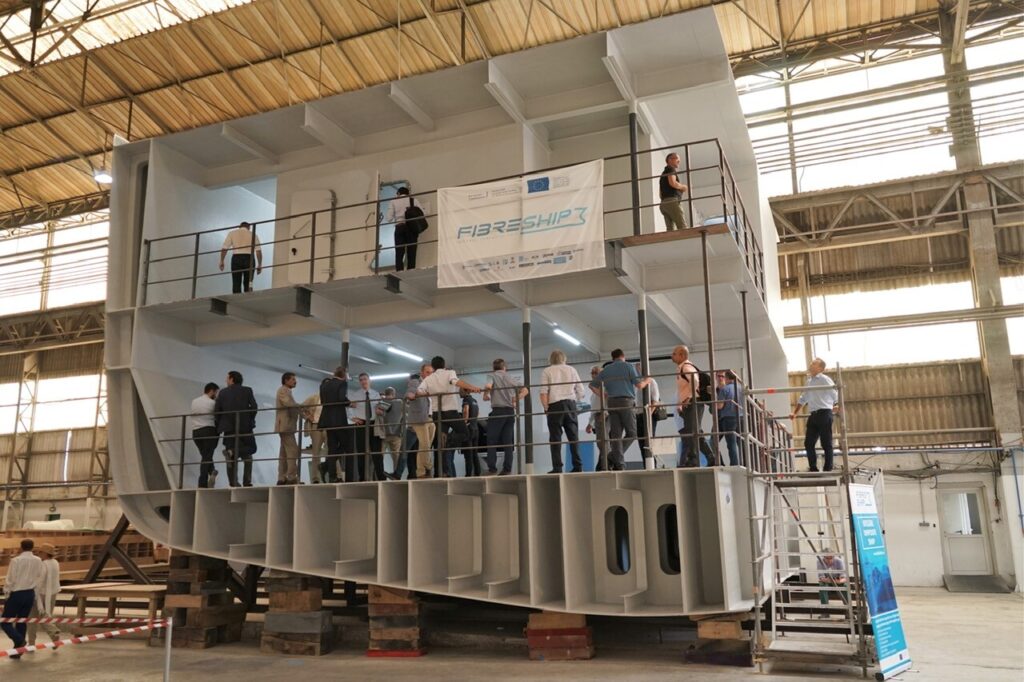 Menschen besichtigen ein großes Schiffsrumpfmodell in einer Industriehalle, mit einem Banner mit der Aufschrift „FIBRESHIP3“.