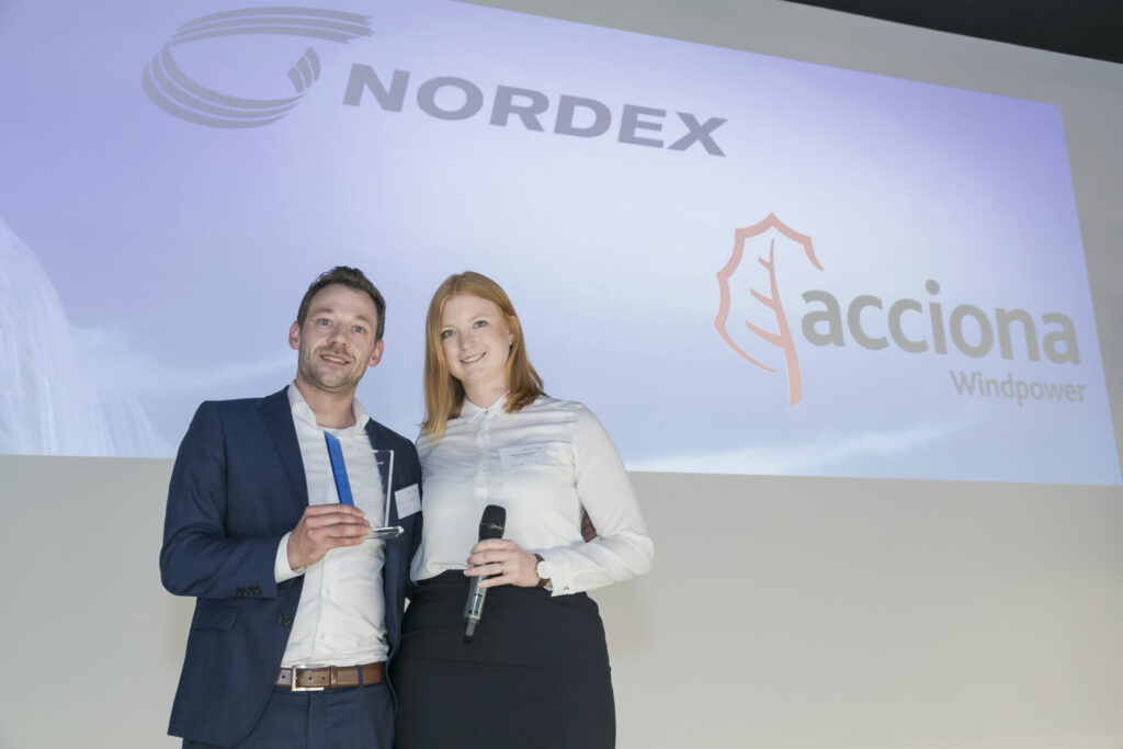 Zwei Personen auf einer Bühne vor einer Projektionswand mit den Logos von Nordex und Acciona Windpower, eine Person hält eine Trophäe, die andere ein Mikrofon.