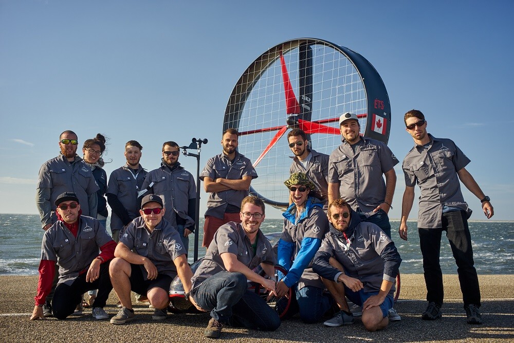 Eine Gruppe von Personen in grauen Hemden steht und kniet vor einem großen Ventilator am Meer.