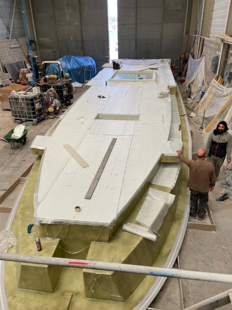 Ein großes Boot oder Schiffsrumpfmodell wird in einer Werkhalle von zwei Männern bearbeitet.