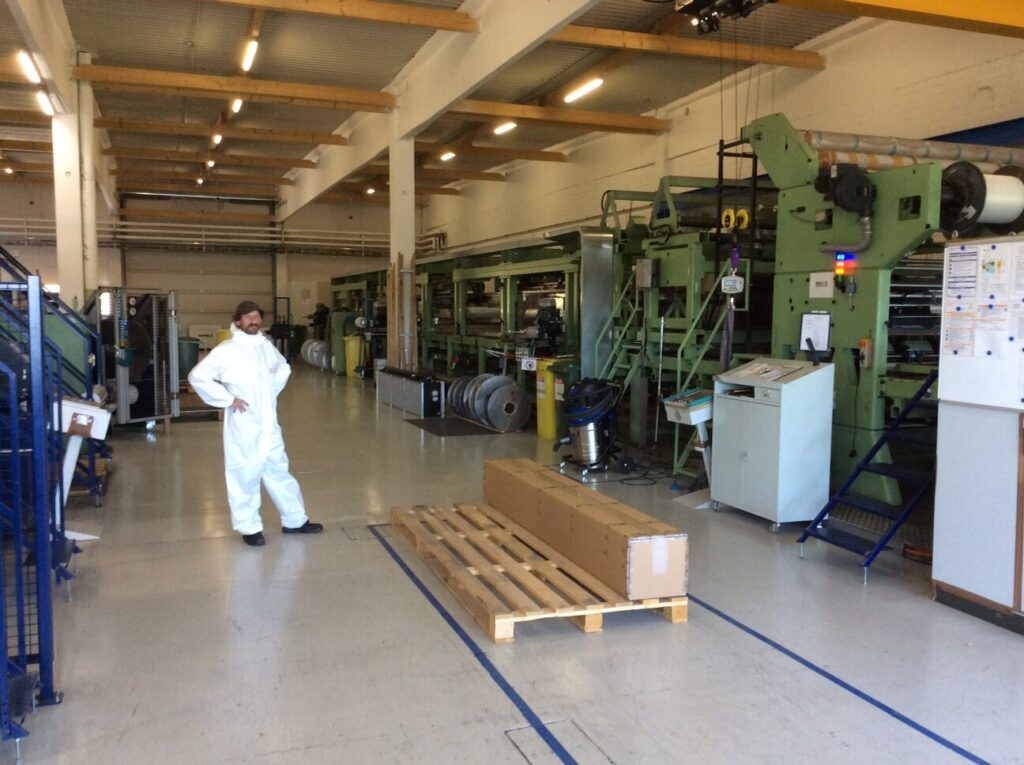 Ein Mann in weißer Schutzkleidung steht in einer großen Industriehalle neben Maschinen und einer Palette mit einem Karton.