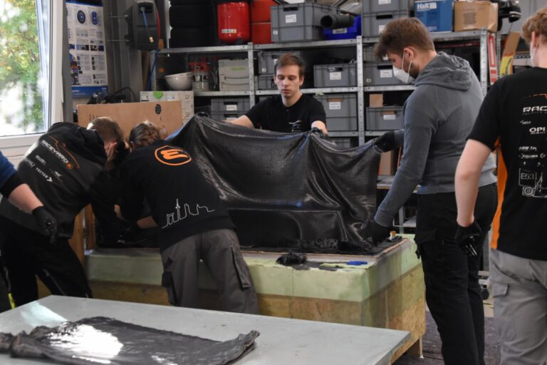 Mehrere Personen in einer Werkstatt halten und bearbeiten ein großes schwarzes, glänzendes Materialstück auf einer Arbeitsfläche.