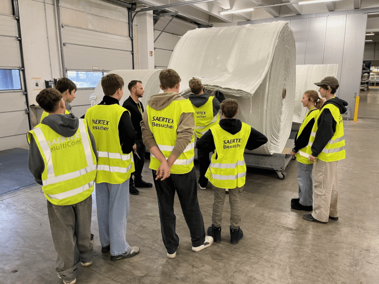 Eine Gruppe von Besuchern in gelben Warnwesten steht in einer Fabrikhalle vor einer großen Papierrolle auf einem Transportwagen.