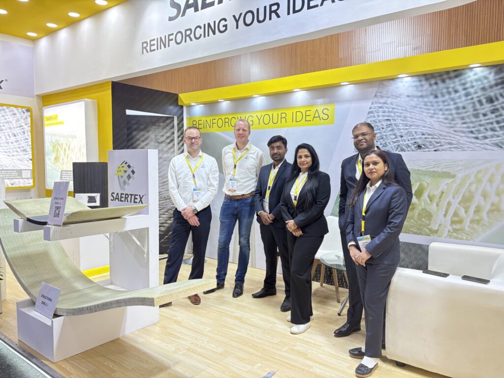 Six people are standing at a SAERTEX booth next to material samples and a large banner that reads „REINFORCING YOUR IDEAS.“.