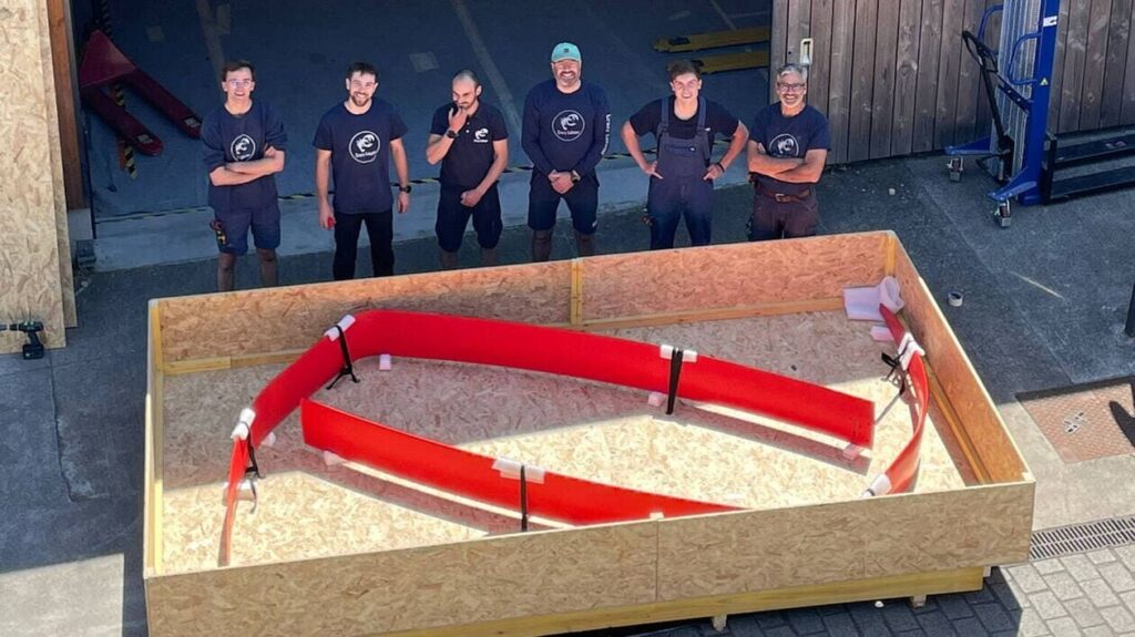 A group of six people in work clothes stand in front of a large wooden frame with a red construction.